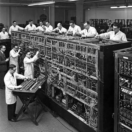 A row of technicians in lab coats working along an enormous early mainframe, patching cables across banks of exposed circuitry