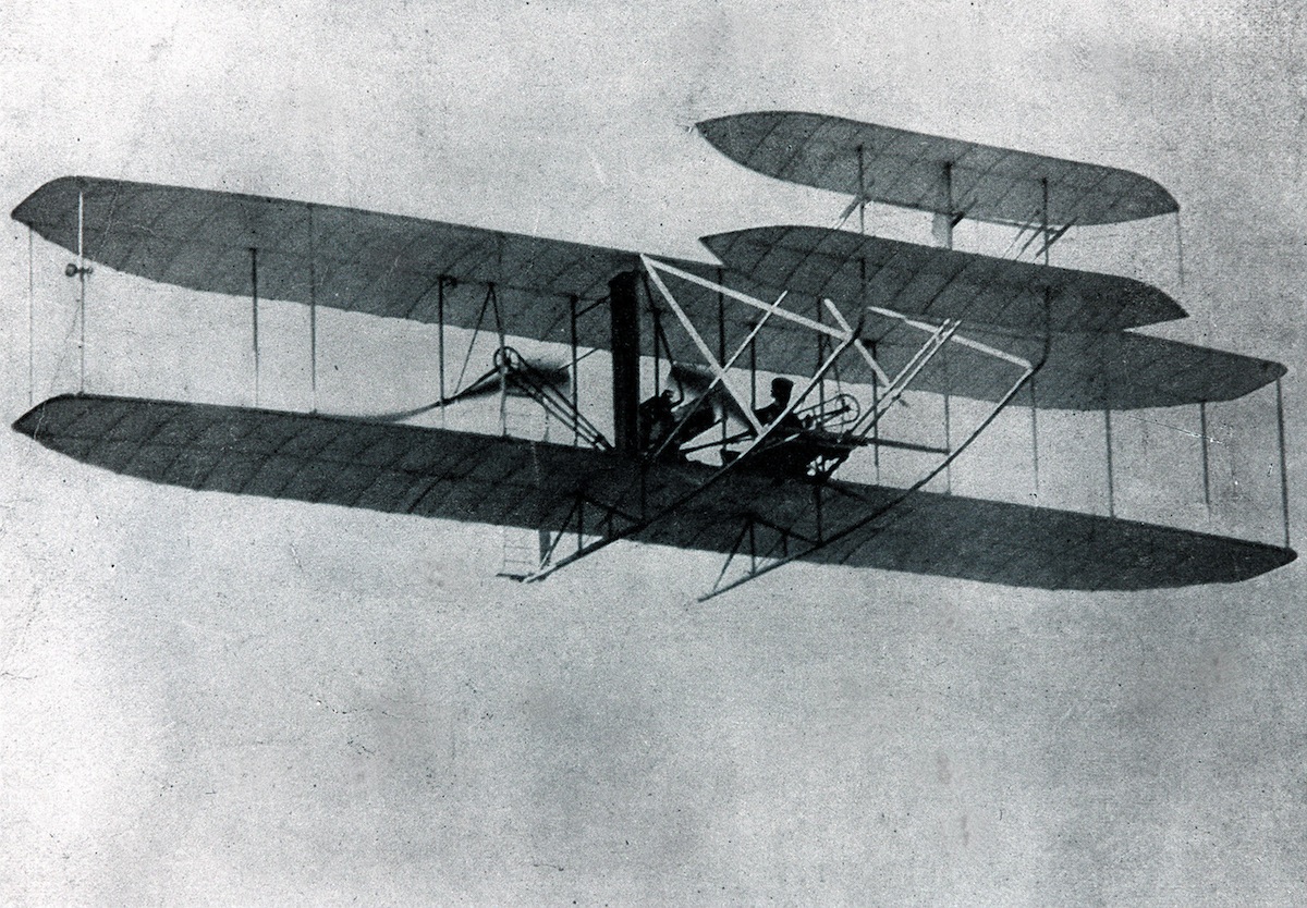 The Wright brothers' biplane in the air, seen from below, a pilot lying prone across the lower wing